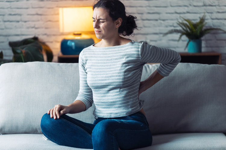 Woman sitting on couch with hand on her back, expressing pain.