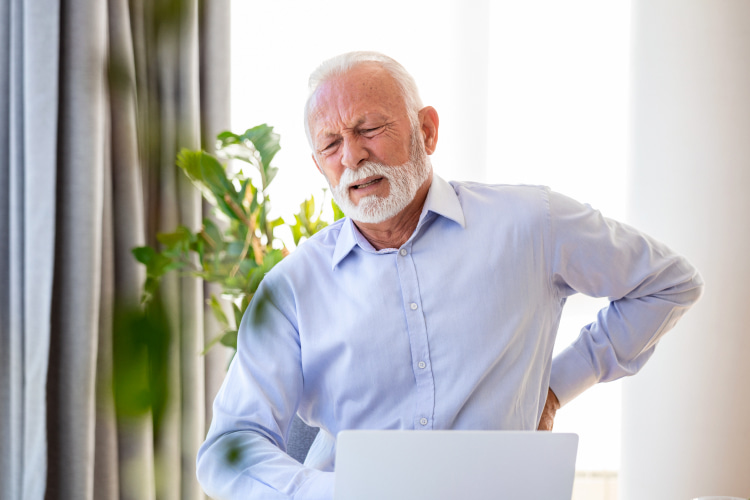 Senior man standing by a laptop, grimacing and holding his lower back in pain.